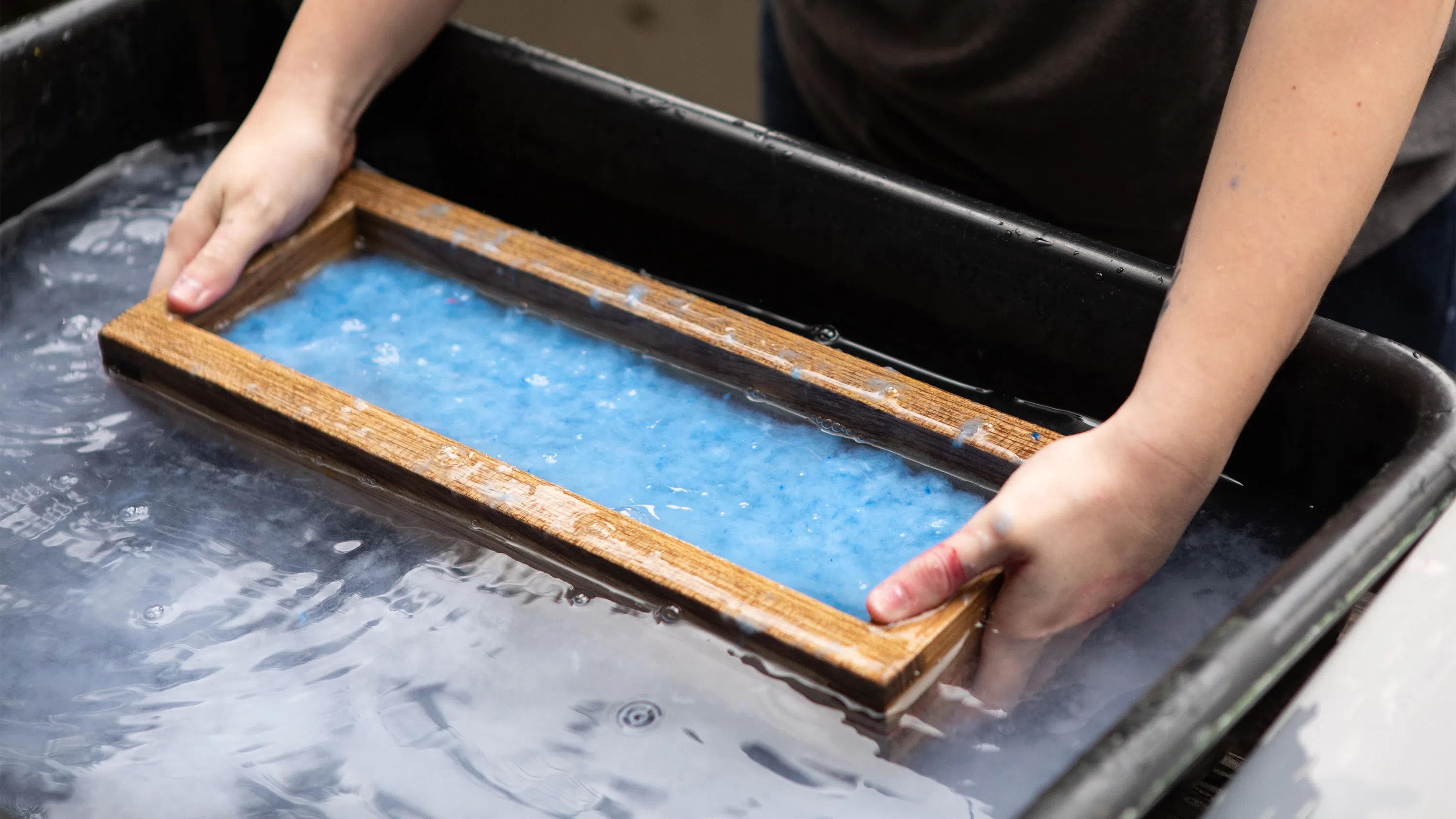 A close-up of hands submerging a screen during the paper-making process.