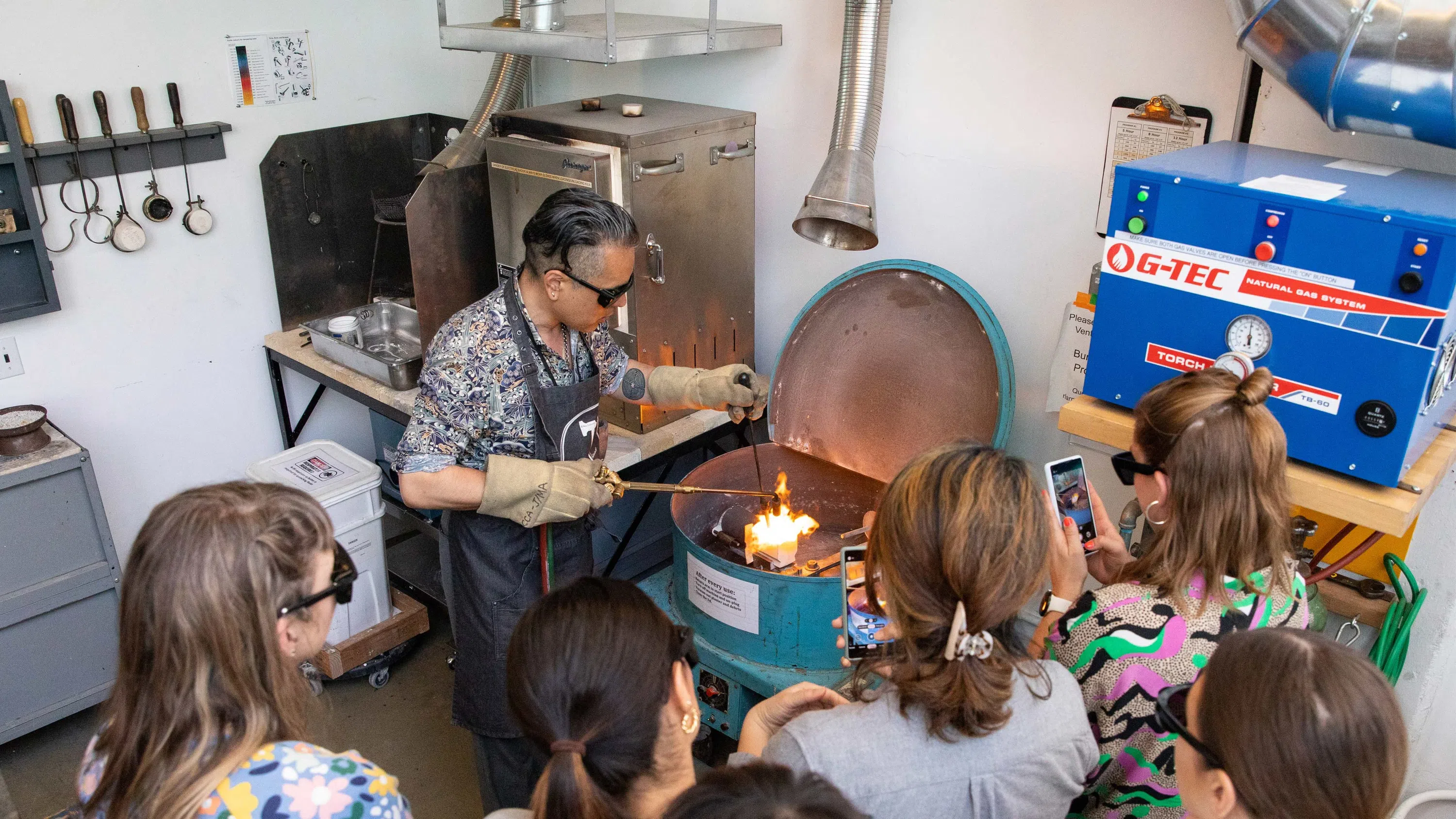An instructor holds a torch over an open kiln in front of a group of people. 