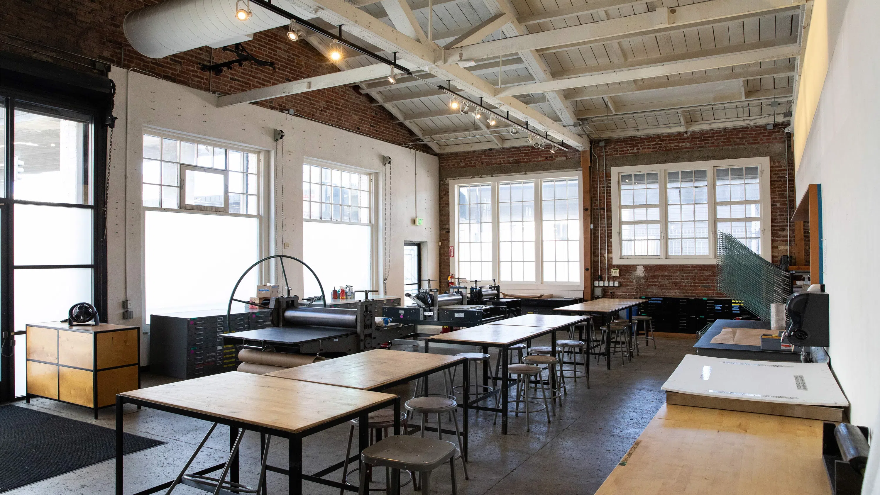 Tables, chairs, and printmedia equipment in a large room with brick walls and large windows. 