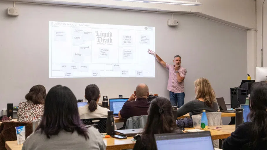 A presenter pointing to a presentation in front of a group of students. 