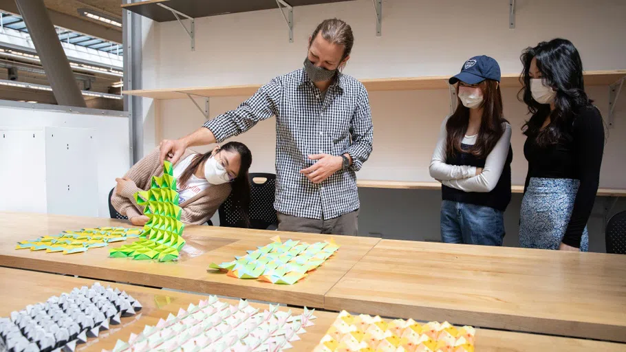 Three students examine three-dimensional woven paper forms in a classroom.