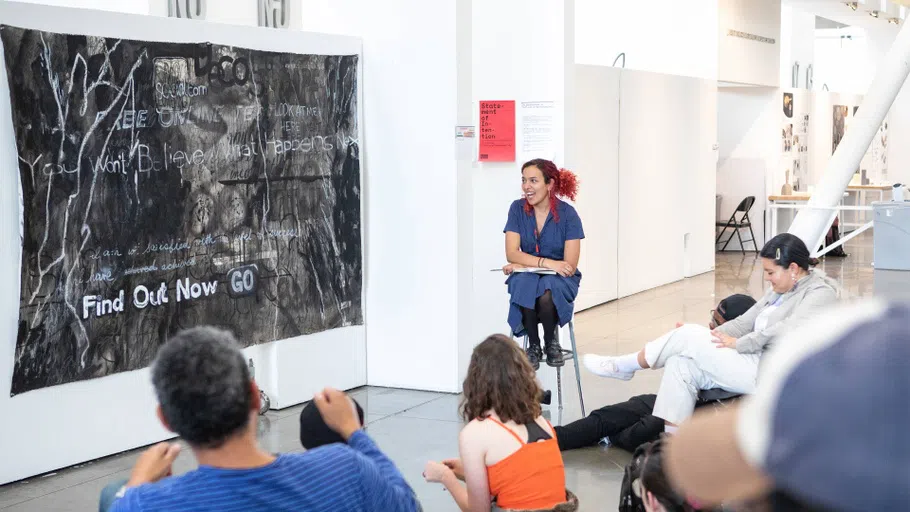 A class critiques a large-scale black painting that includes a line drawing of an animal and text like “Find Out Now,” “Go,” and “You Won’t Believe.”