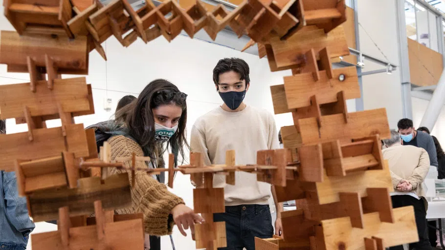 Framed within the model, two students examine a bridge-like structure composed of interlocking wooden pieces.