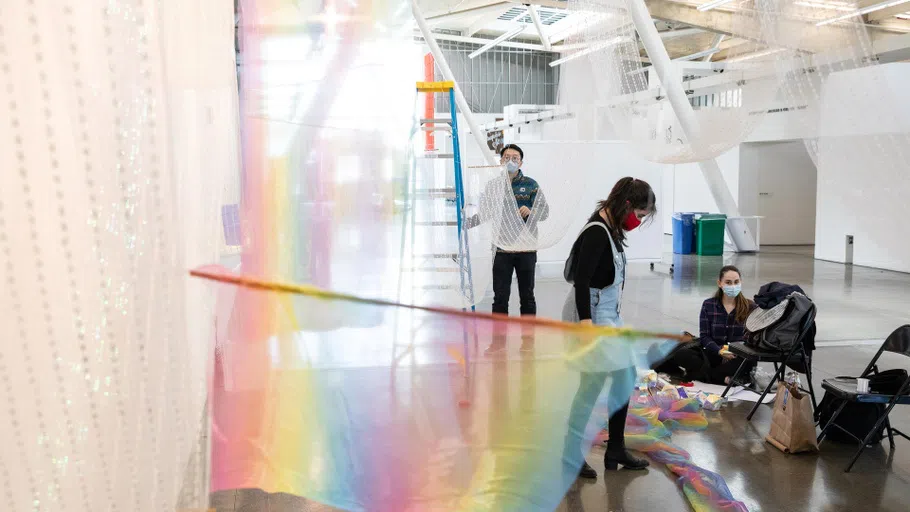 A person stands on a ladder, hanging an installation of gauzy mesh and prisms. Toward the back of the light-filled space, four people suspend large-scale photographs.