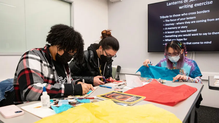 Three students work on cutting and collaging materials; a screen behind describes the related writing exercise.