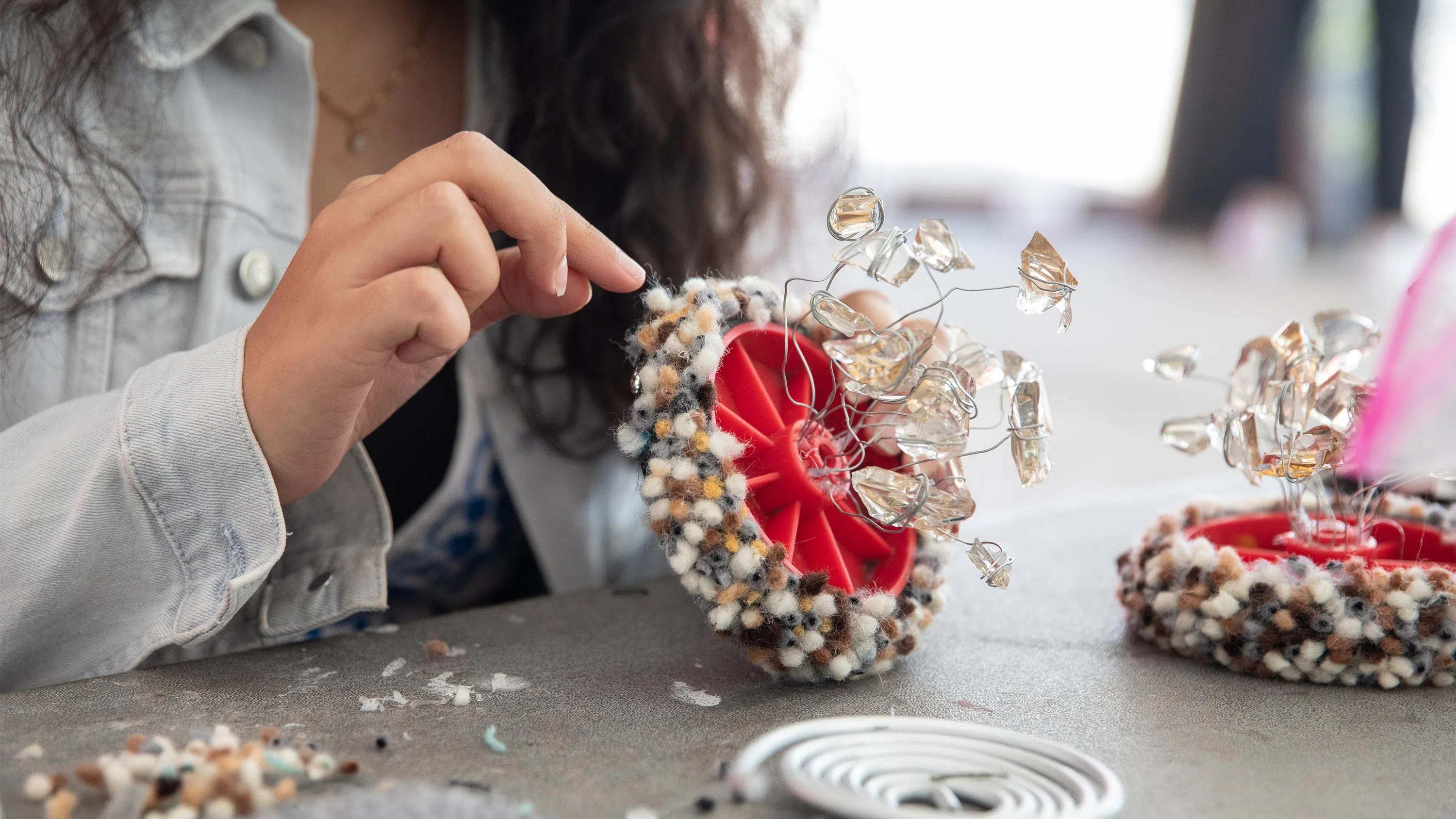 A close-up view of a student working on a flower-like sculpture made of wire, beads, and plastic.
