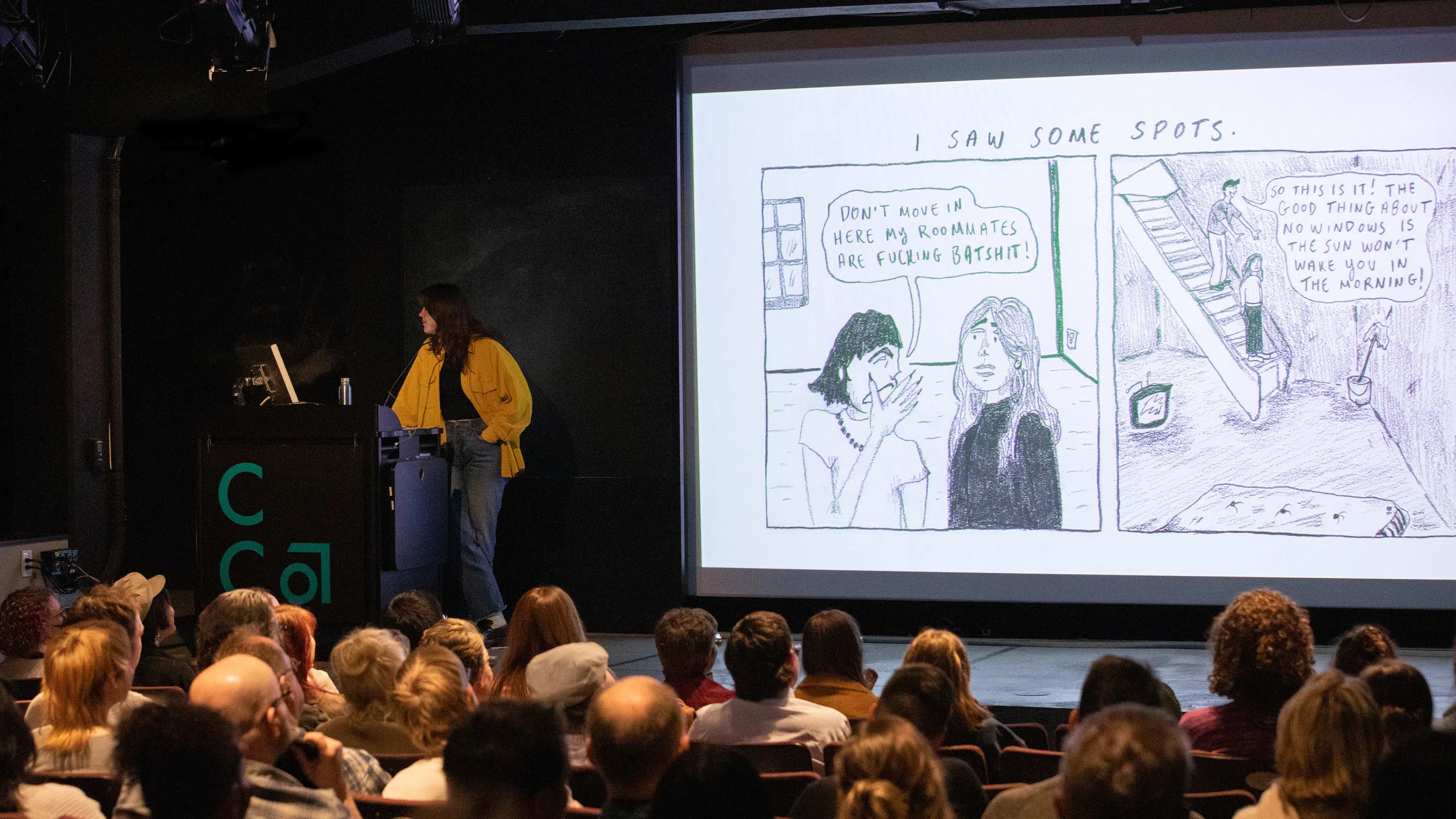 A student stands on a stage projecting a comic strip onto a screen. 