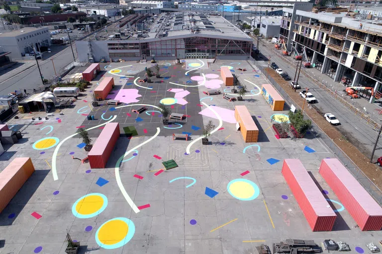 An aerial view of CCA's backlot with colorful chalk drawings of abstract shapes, painted containers, potted plants, and picnic tables decorating the concrete area. The main building recedes in the background.