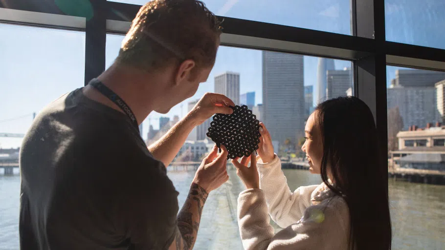 Two students hold up a black, porous 3D-printed sample against a window overlooking the Embarcadero.