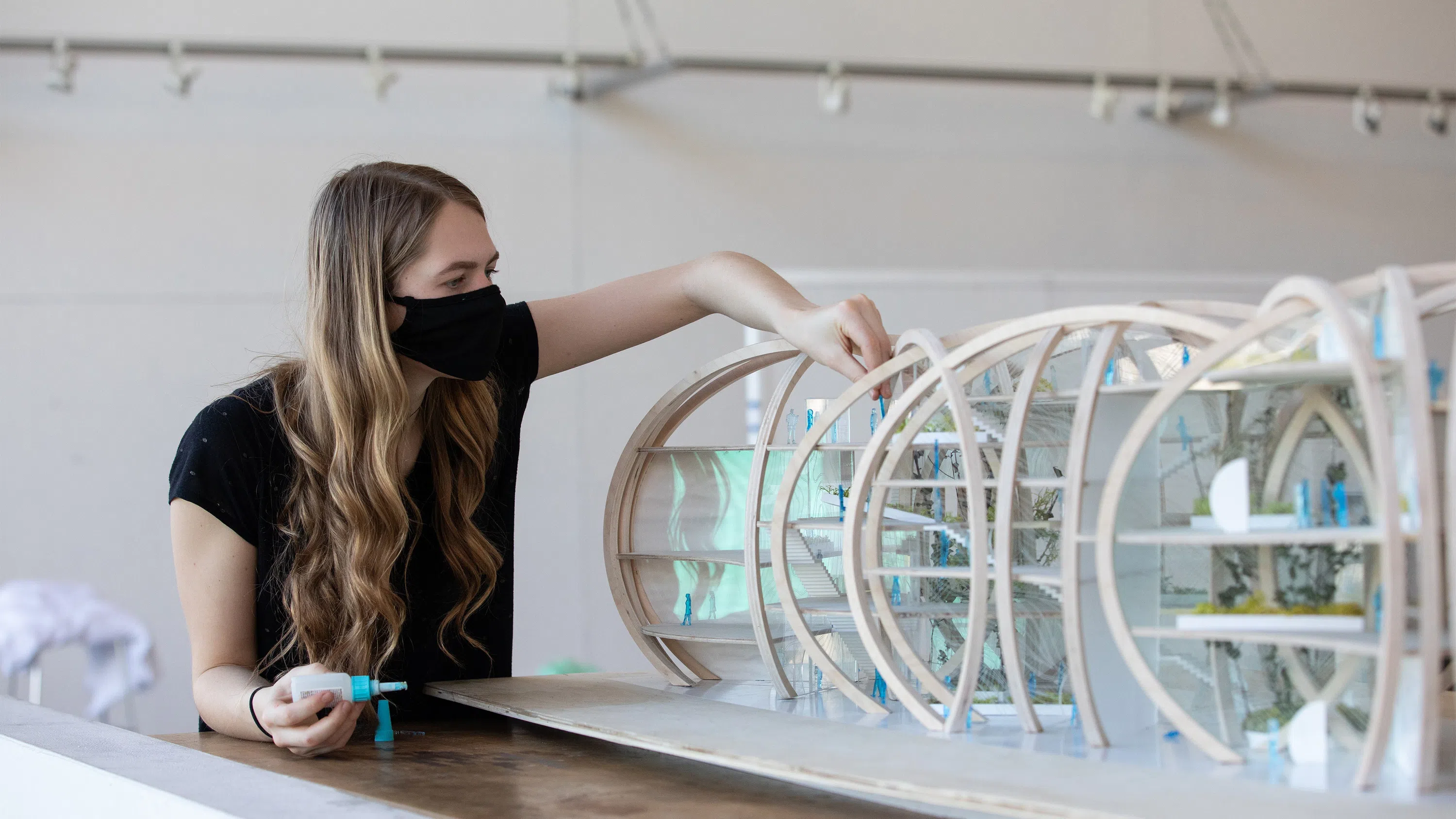 A young woman superglues a plexiglass and balsa wood architectural model.