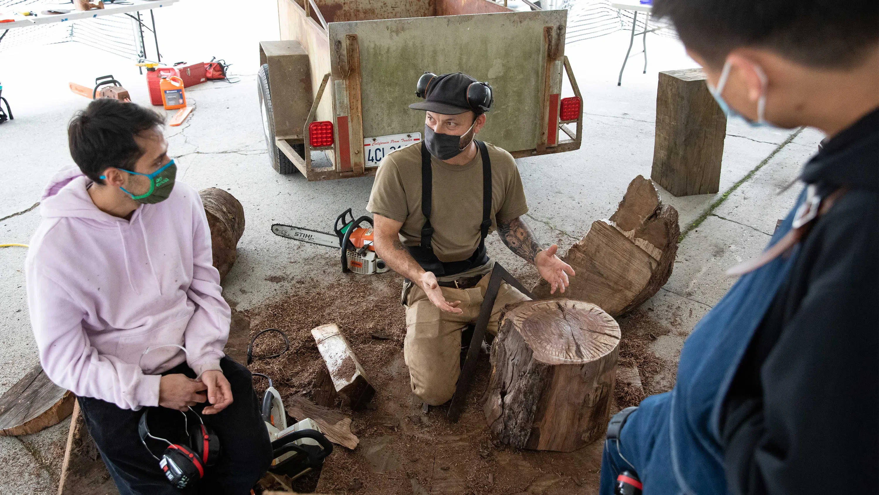 Ido Yoshimoto kneels to demonstrate chainsaw techniques on a wooden stump for two students on the Backlot.