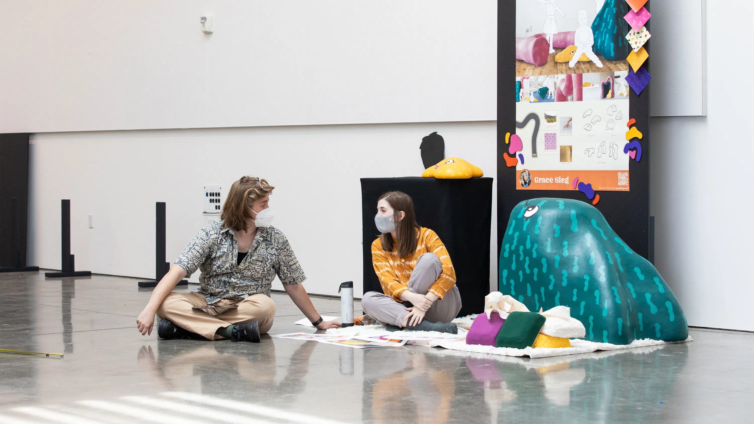 Two students sit on the floor with large-scale, colorful objects and pillows; a pin-up board behind them includes documentation and inspiration for the project, titled Playwell.