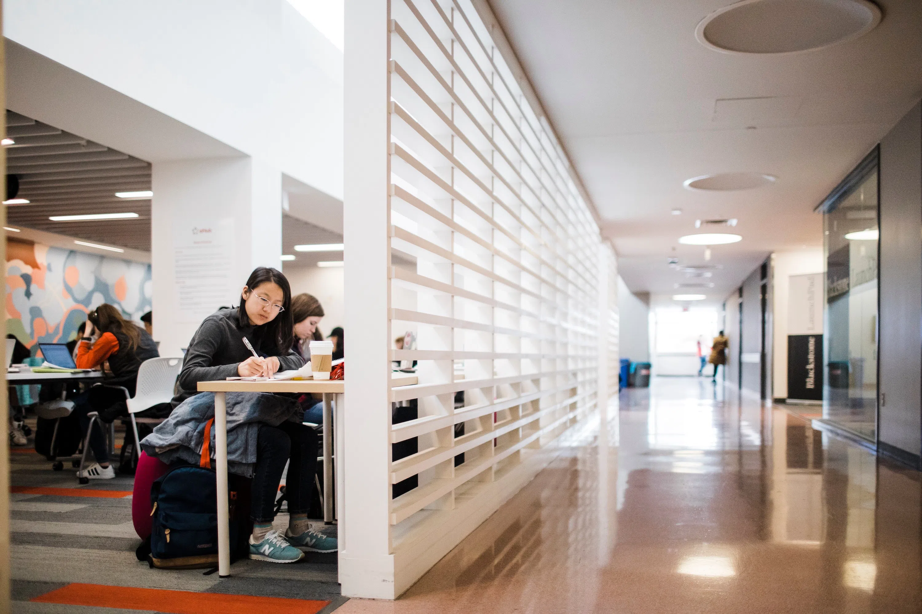 On the left, students sit a table writing and typing on their computers. On the right, a long hallway lined with offices.