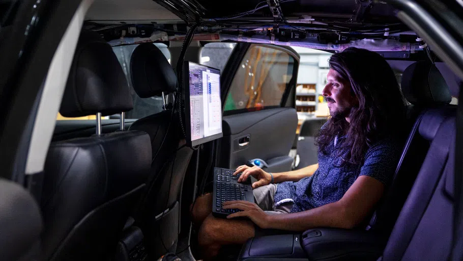 A man with a beard and long hair sits in the back seat of a car. In front of him is a bright screen and a keyboard on which he is typing.