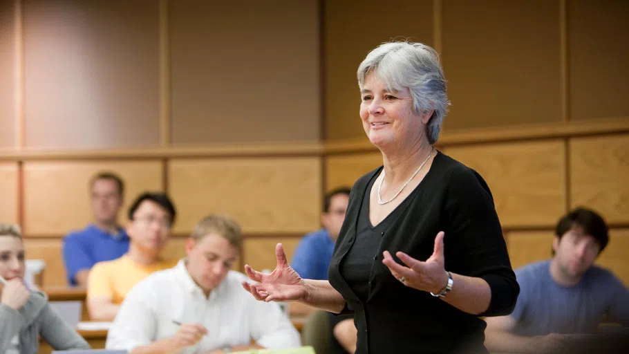 A faculty member reaching out her hands while teaching a class. A handful of students are seen in the background, sitting in a long row.