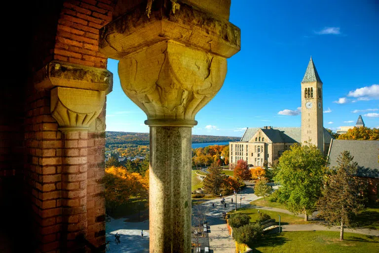 A view of McGraw Tower, Uris Library and Ho Plaza on a sunny autumn day with Cayuga Lake in the distance, taken from Barnes Hall with red brick and a marble column at the front left of the image.