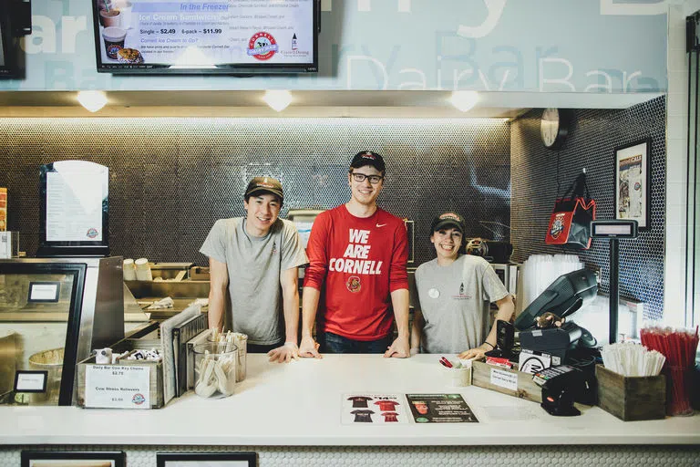 Three student workers smile behind the counter at the Cornell Dairy Bar at Stocking Hall.