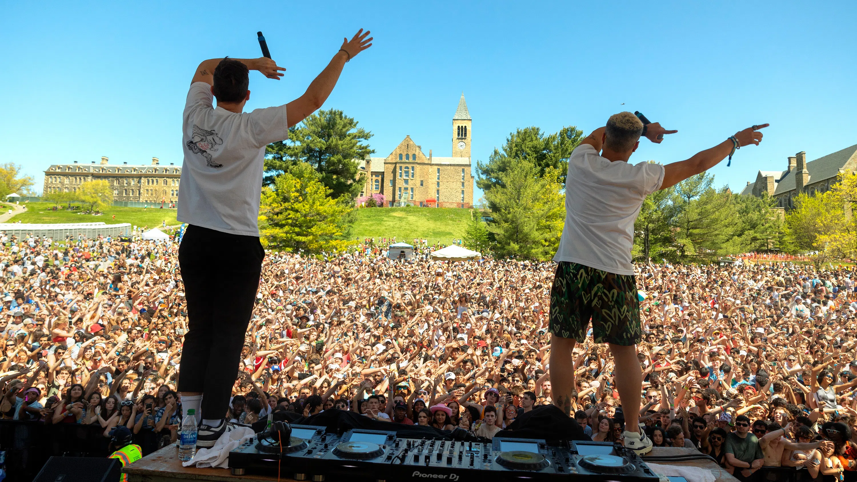 Two performers at Slope Day. Photo is taken from behind them on the stage, looking out toward an audience of thousands of Cornell students. Trees and McGraw Tower are in the background.