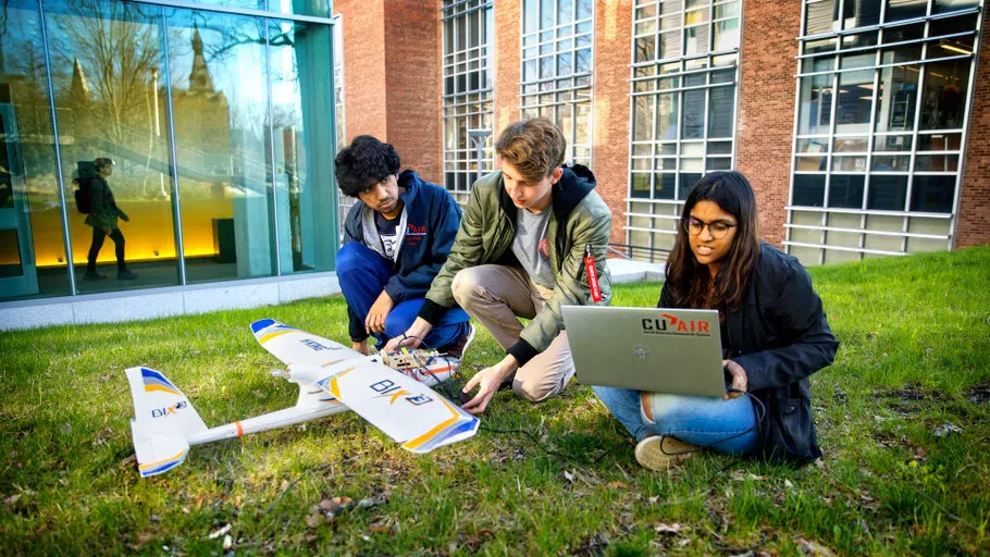 Three students kneel on the ground as they work on a prototype of a small, unmanned aerial unit.