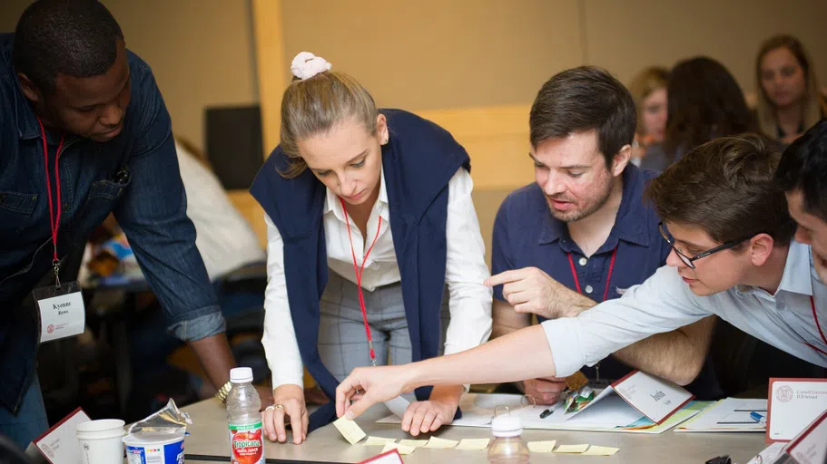 Four college students, each wearing a lanyard and name tag, look down at a table at a set of small Post-It Notes.