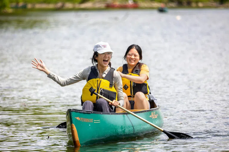 Two students wearing yellow life vests and big smiles row with oars on Beebe Lake in a green canoe during Senior Days.