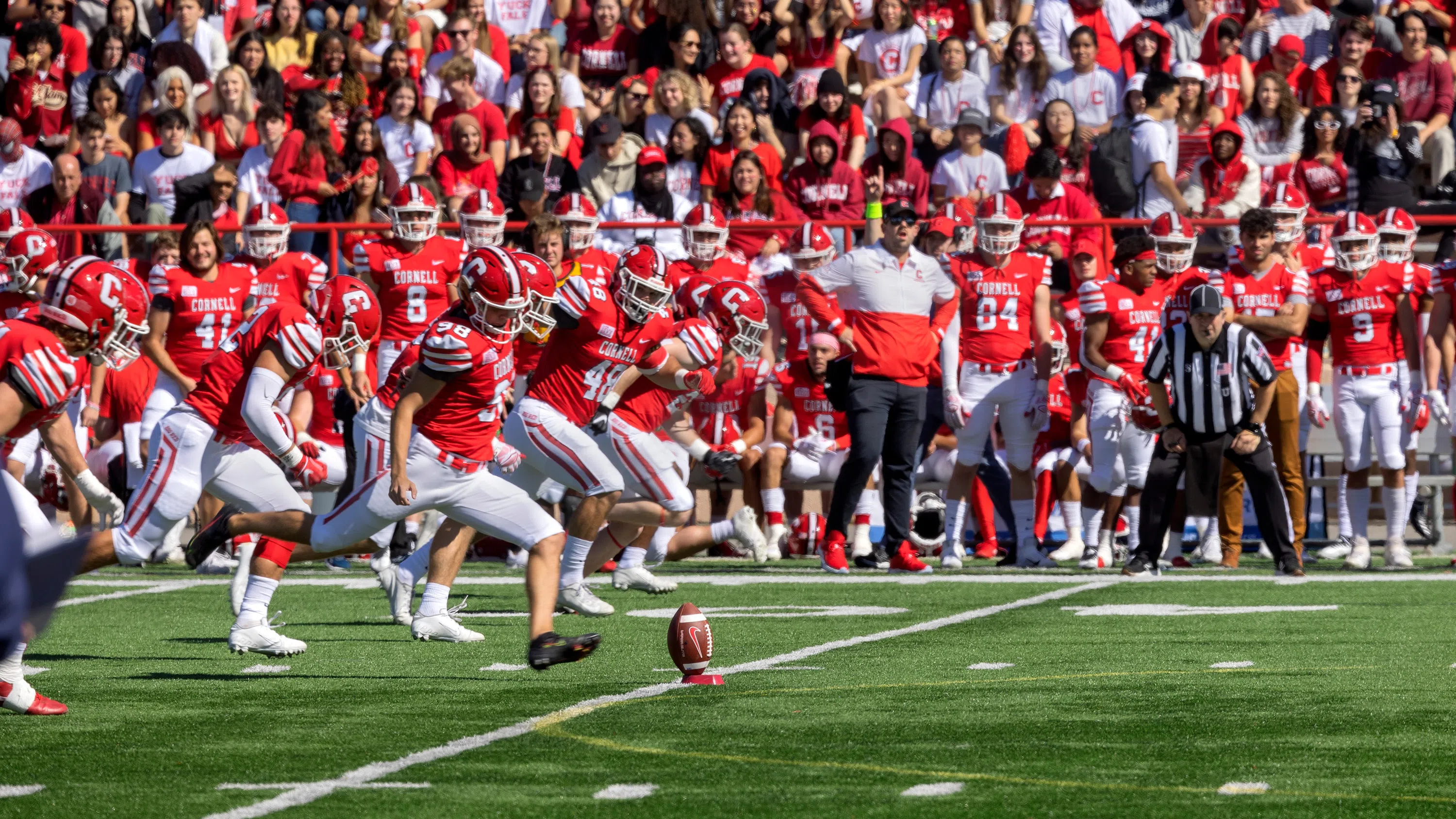 A line of Cornell football players advances toward a football placed on a small red stand during a game.