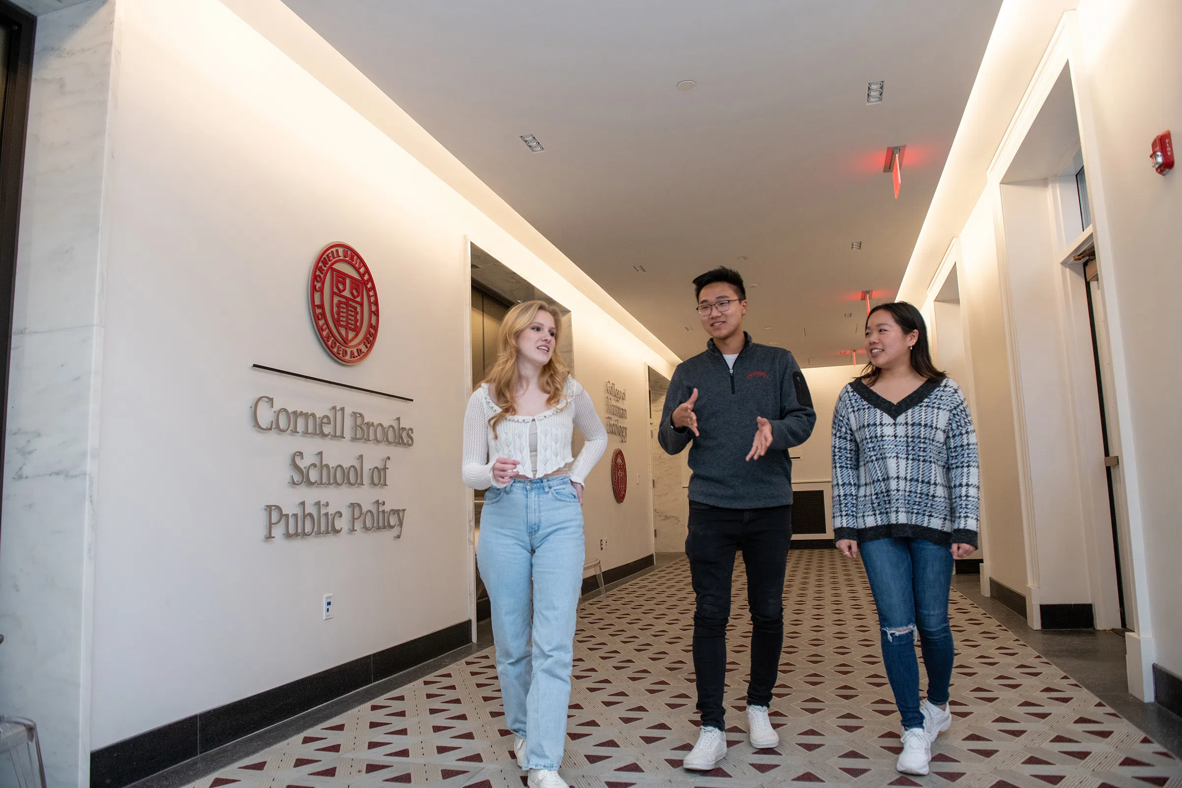 Three students walk in a hallway, talking. On the wall to the left is a Cornell University seal and raised lettering that reads “Cornell Brooks School of Public Policy.”