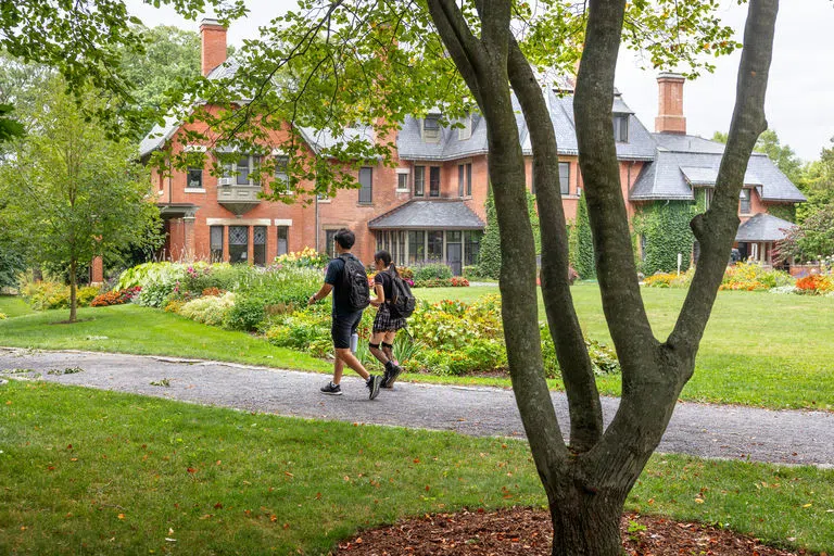 Students walk along a path beside colorful gardens in full bloom with the A.D. White House in the distance.