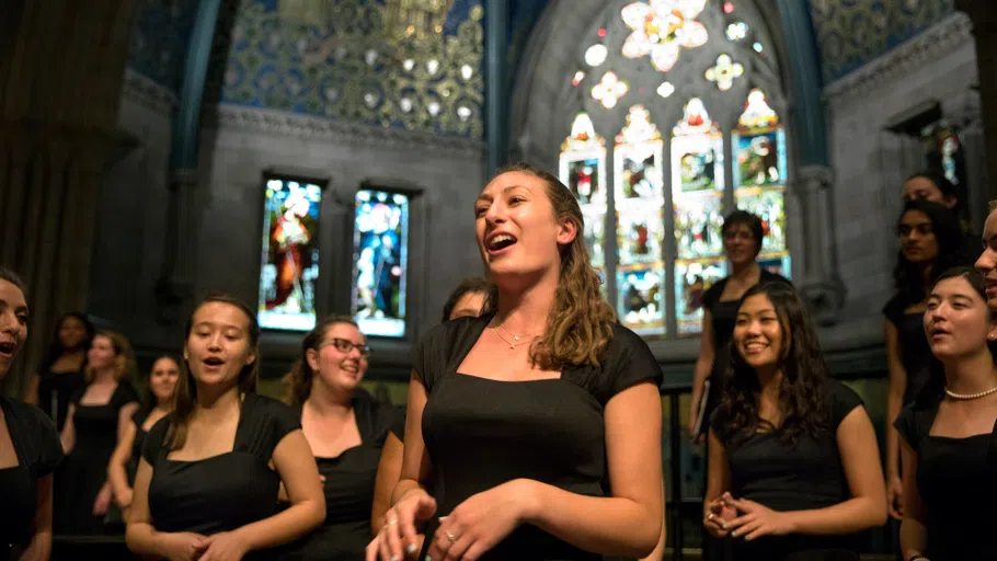 Choir singers perform in Sage Chapel, with multiple stained-glass windows behind them.