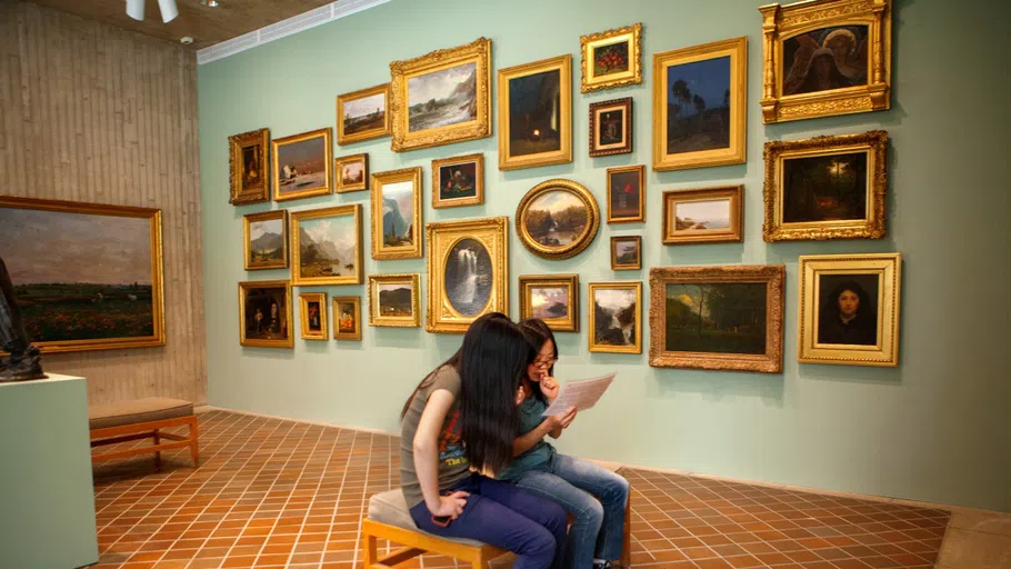 Two visitors sit on a bench at the Johnson Museum surrounded by a gallery of small paintings mounted on the walls.