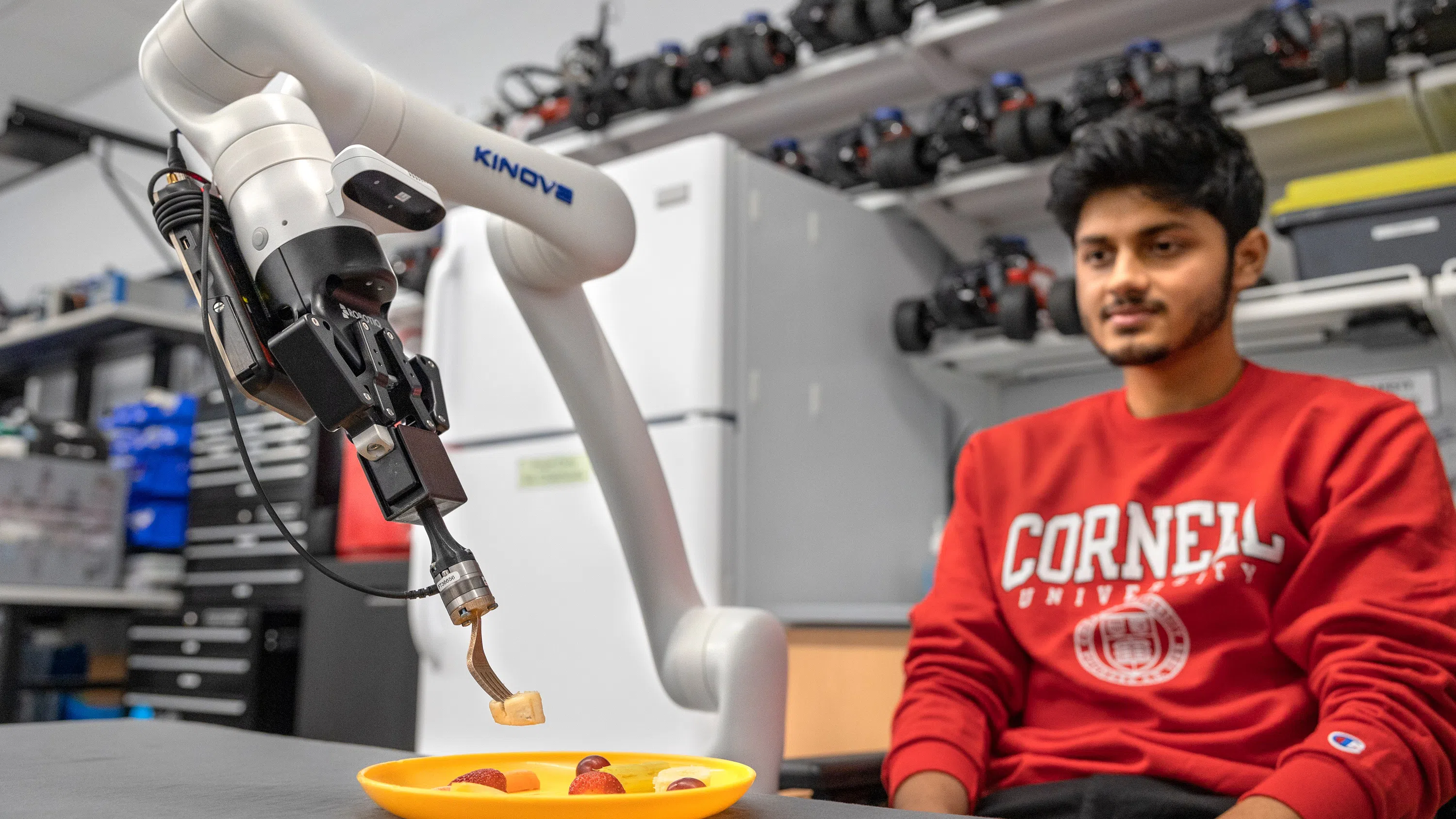 A student, right, wearing a red Cornell sweatshirt, watches a robot arm that is holding a fork with a piece of banana.