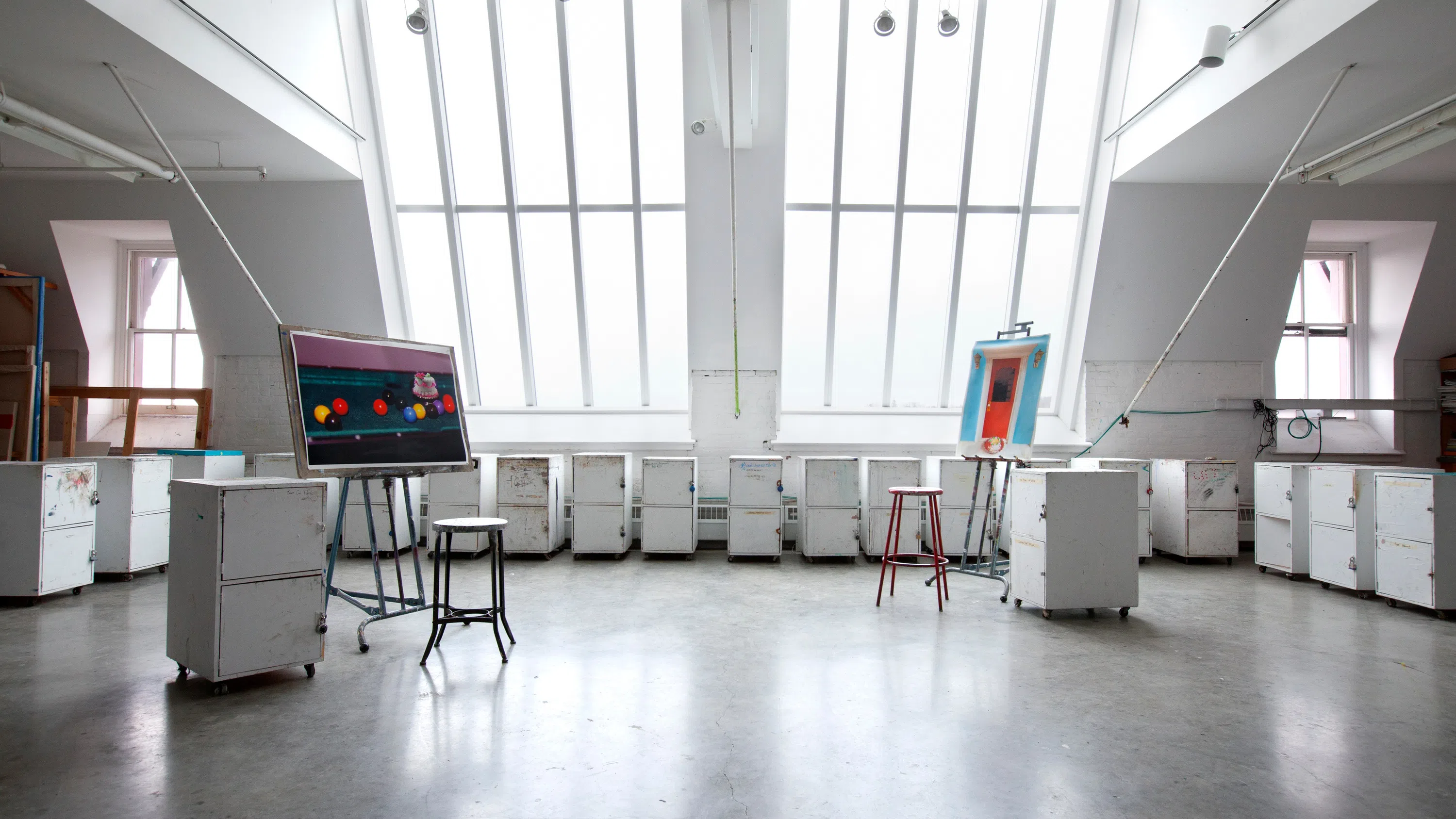 A wide-open studio space with very large sunlight windows and several easels with nearly finished paintings on them. There are more than a dozen portable art cabinets - white but with lots of markings on them.