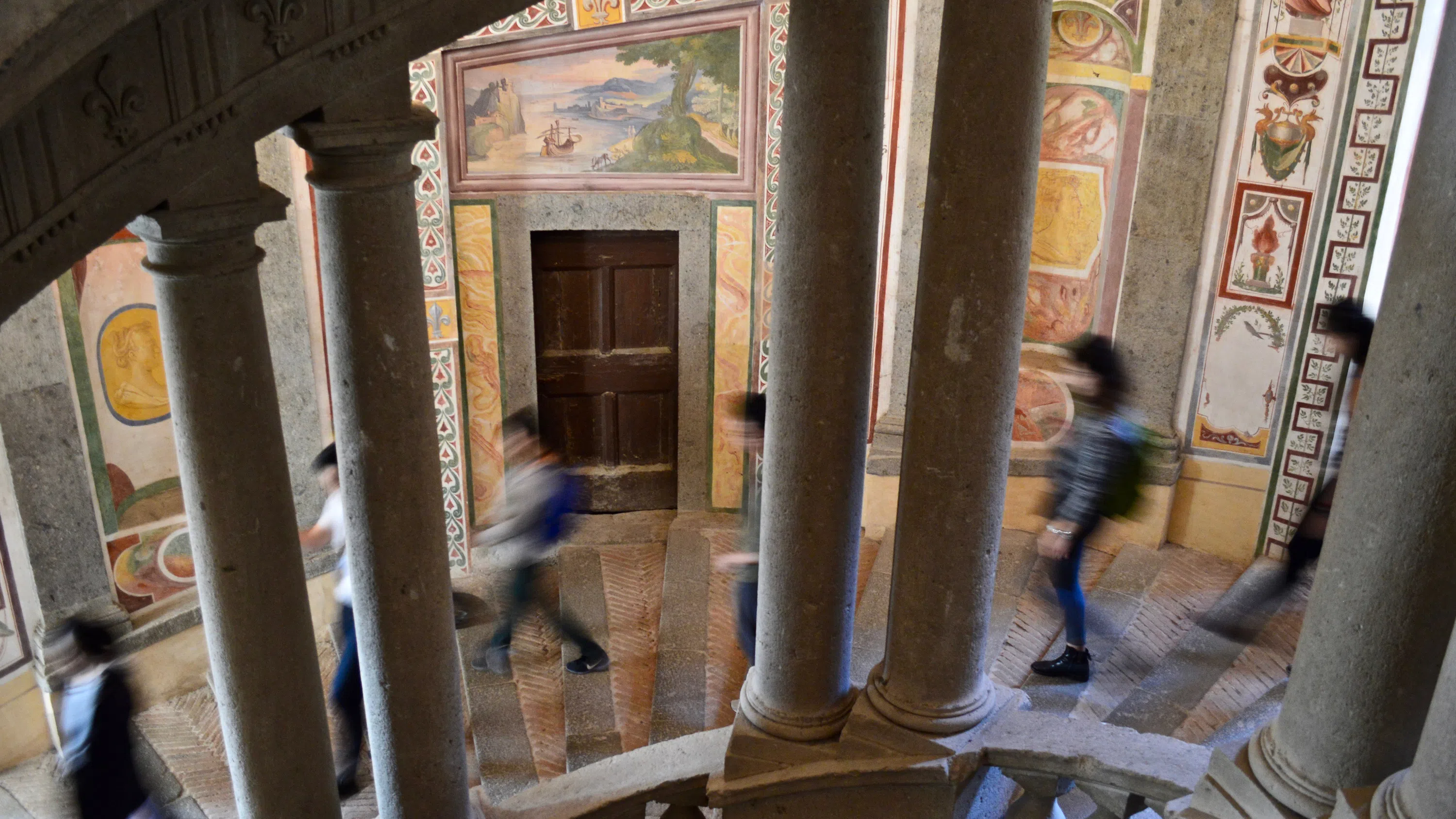 Students (who are out of focus) descend a spiral staircase with stone railings and beautiful inlay artwork on the wall.