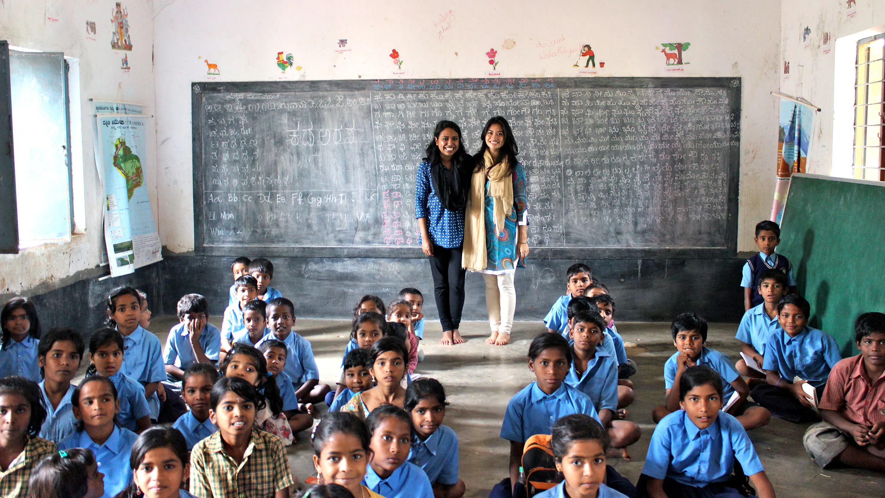 Two women stand in a classroom in India. In front of them are about three dozen schoolchildren, all sitting on the floor and most wearing blue shirts. Behind the women is a chalkboard covered in writing.