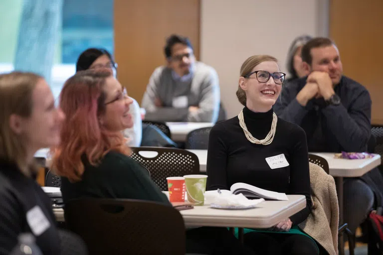 Two people sit at a table with others and smile, listening to a speaker (not pictured) during the Pathways to Success Symposium, an annual professional development event at the Graduate School.