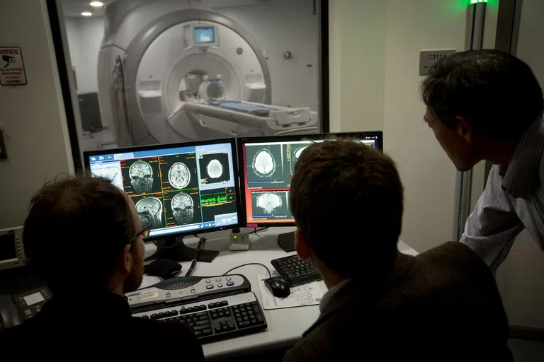 Three people, seen from behind in silhouette, look at images of brain scans on monitors in front of them. In the background is an MRI machine.