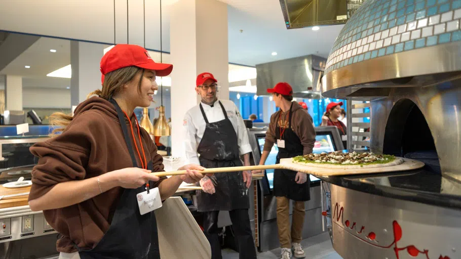 A student puts a pizza on a wooden board into a large oven in a dining hall kitchen. 