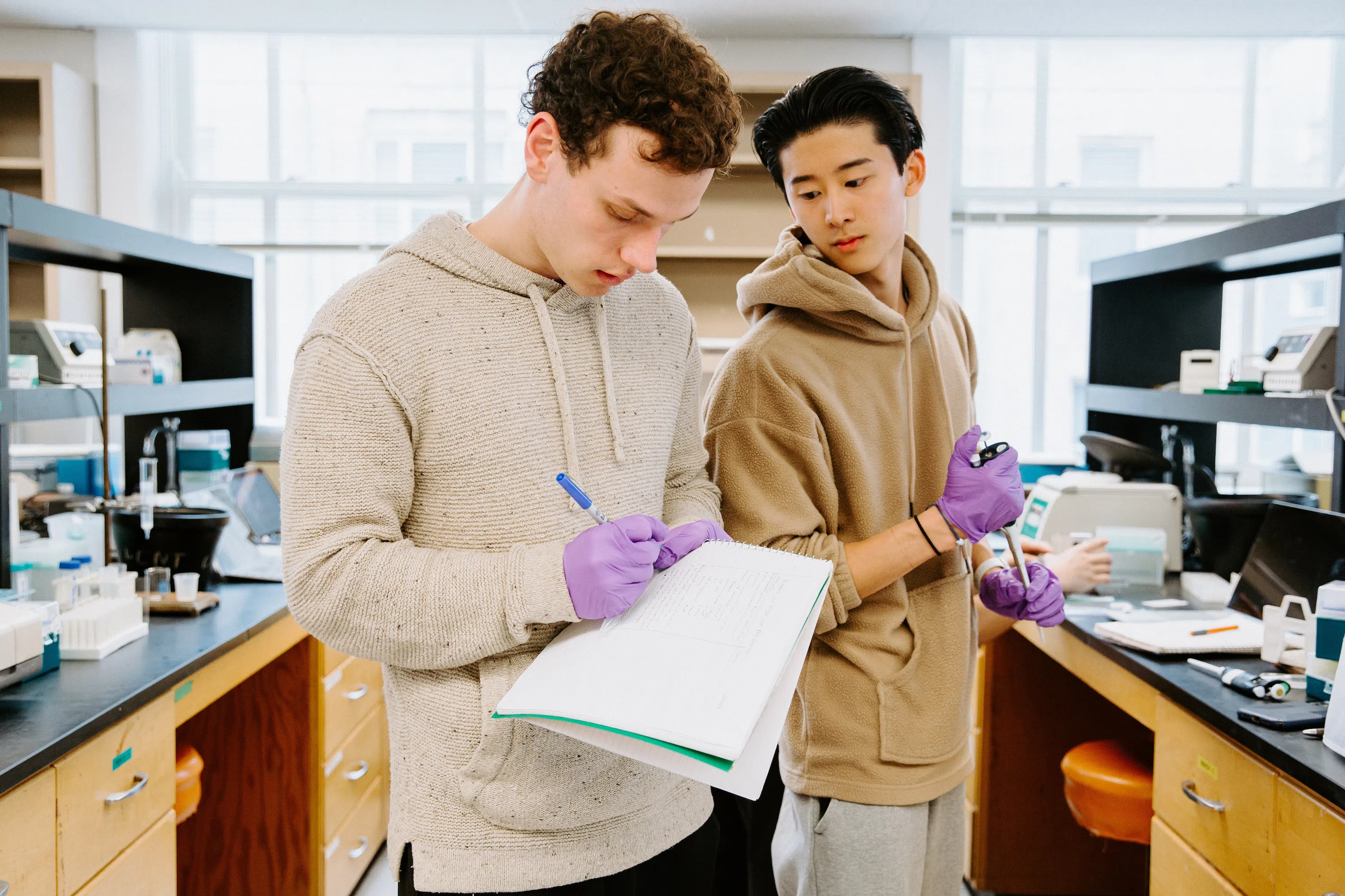 Two students, both wearing hoodie sweatshirts and purple gloves, work on a lab experiment. The student at left is taking notes while the other student looks down at the notebook.