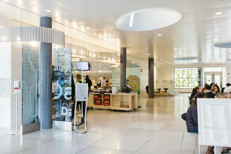 A bright view of the Cornell Dairy Bar from the entrance of Stocking Hall with a seating area to the right and a sign, menu and cash register to the left.