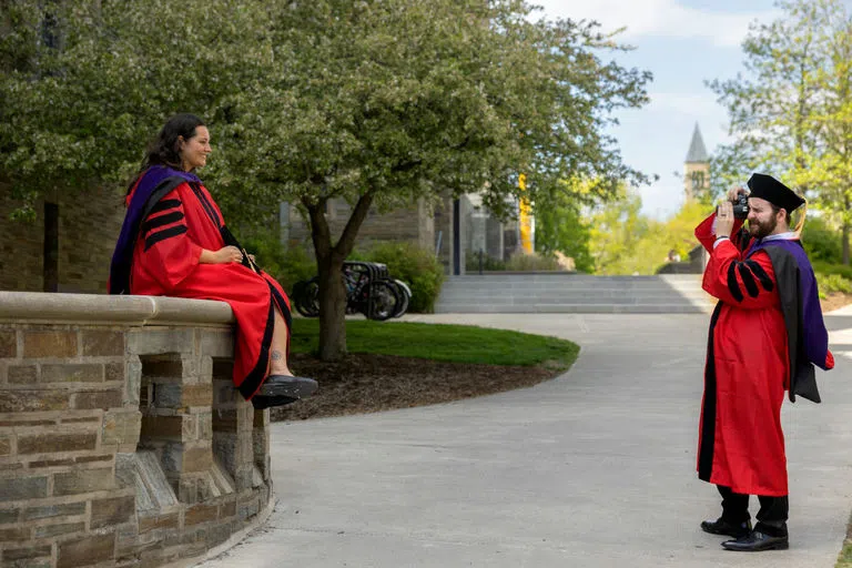 Students wearing red robes and regalia take graduation photos in front of Myron Taylor Hall.