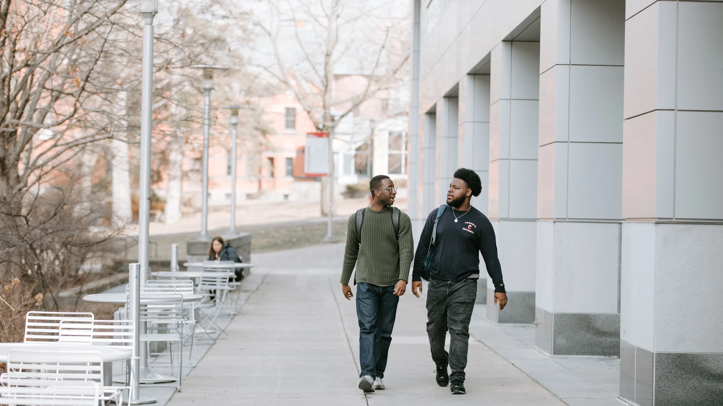 Two students walk past Duffield Hall at Cornell University. To the left are silver metal outdoor tables, with a tall heat lamp at each. To the right are some of the building’s support pillars.