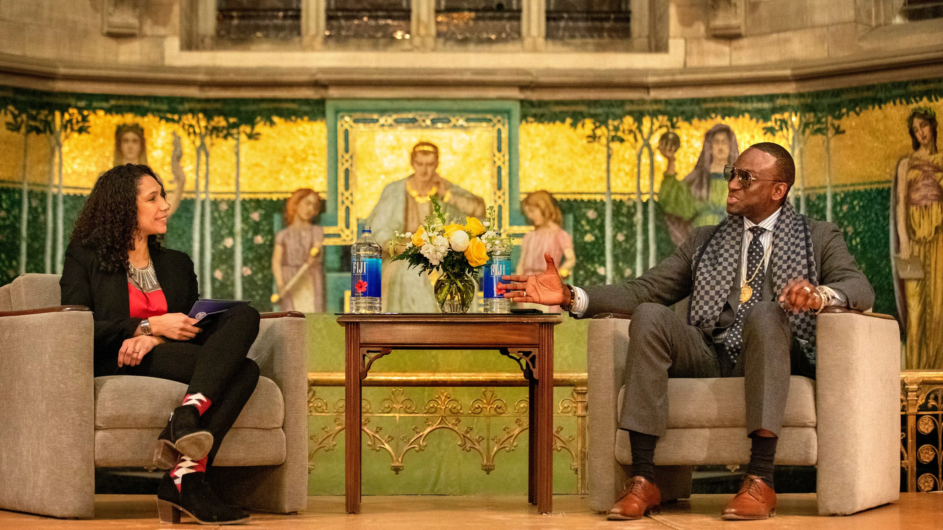 A moderator, left, holds a discussion in Sage Chapel with a guest speaker, right. Between them is a small table with a vase of roses, and behind them is a religious fresco. 