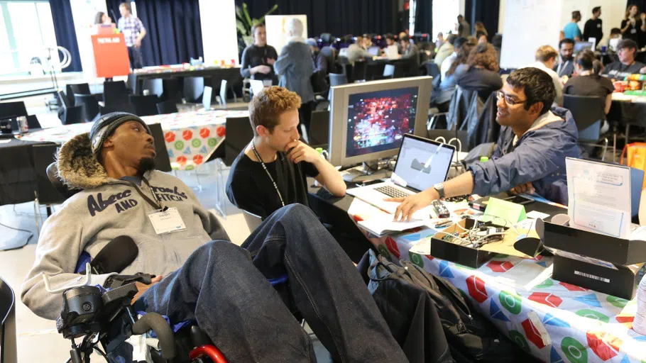 Two individuals, one in a wheelchair and the other seated, speak with another person sitting at a table across from them. On the table is a laptop, a computer monitor and several small boxes of equipment. In the background are several more long tables with similar discussions occurring.