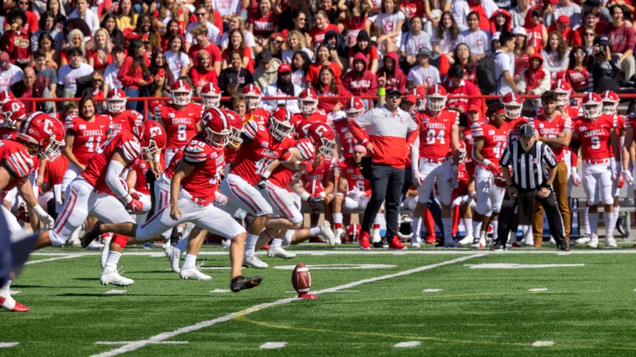 A line of Cornell football players advances toward a football placed on a small red stand during a game.