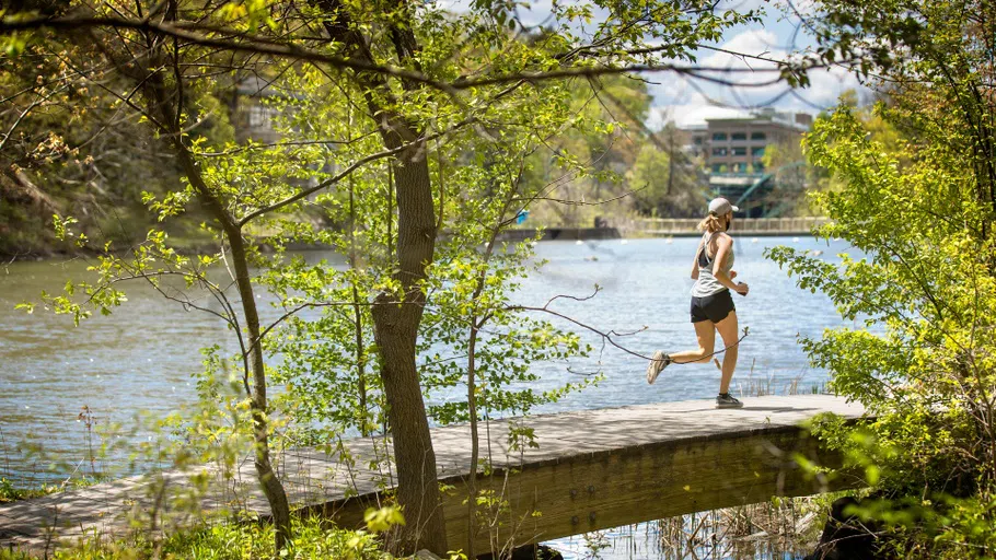 A runner crosses a footbridge over Beebe Lake on a sunny day, surrounded by trees and with Rand Hall in the distance.