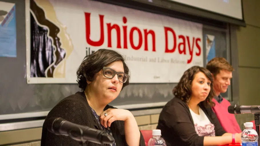 In the foreground at left, a panelist wearing glasses and holding a pen listens to someone speaking off-camera. Above is a banner that reads “Union Days” and to the right are two other panelists, also listening.