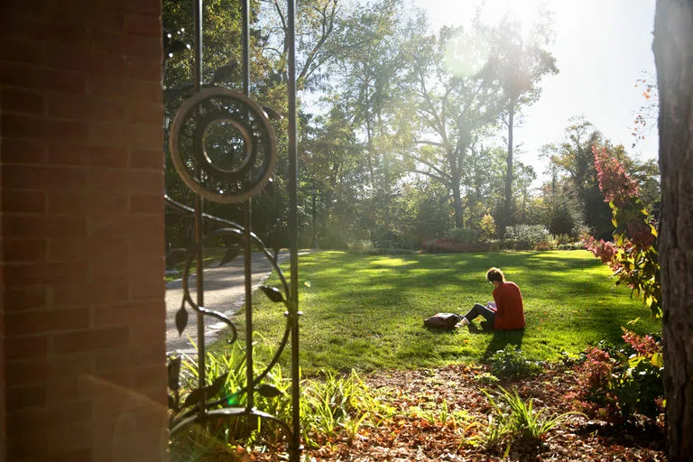 A view taken from the gate of the A.D. White House of a student studying on the lawn dappled with sunlight among the gardens.