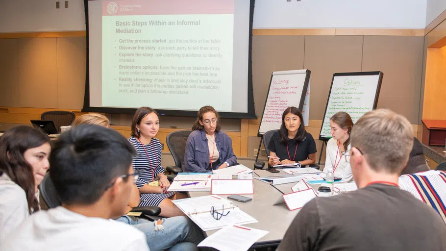 Eight college students sit around a circular table, with binders in front of them. In the background are two easels with writing, as well as a projection screen showing a PowerPoint slide titled, “Basic Steps Within an Informal Mediation.”