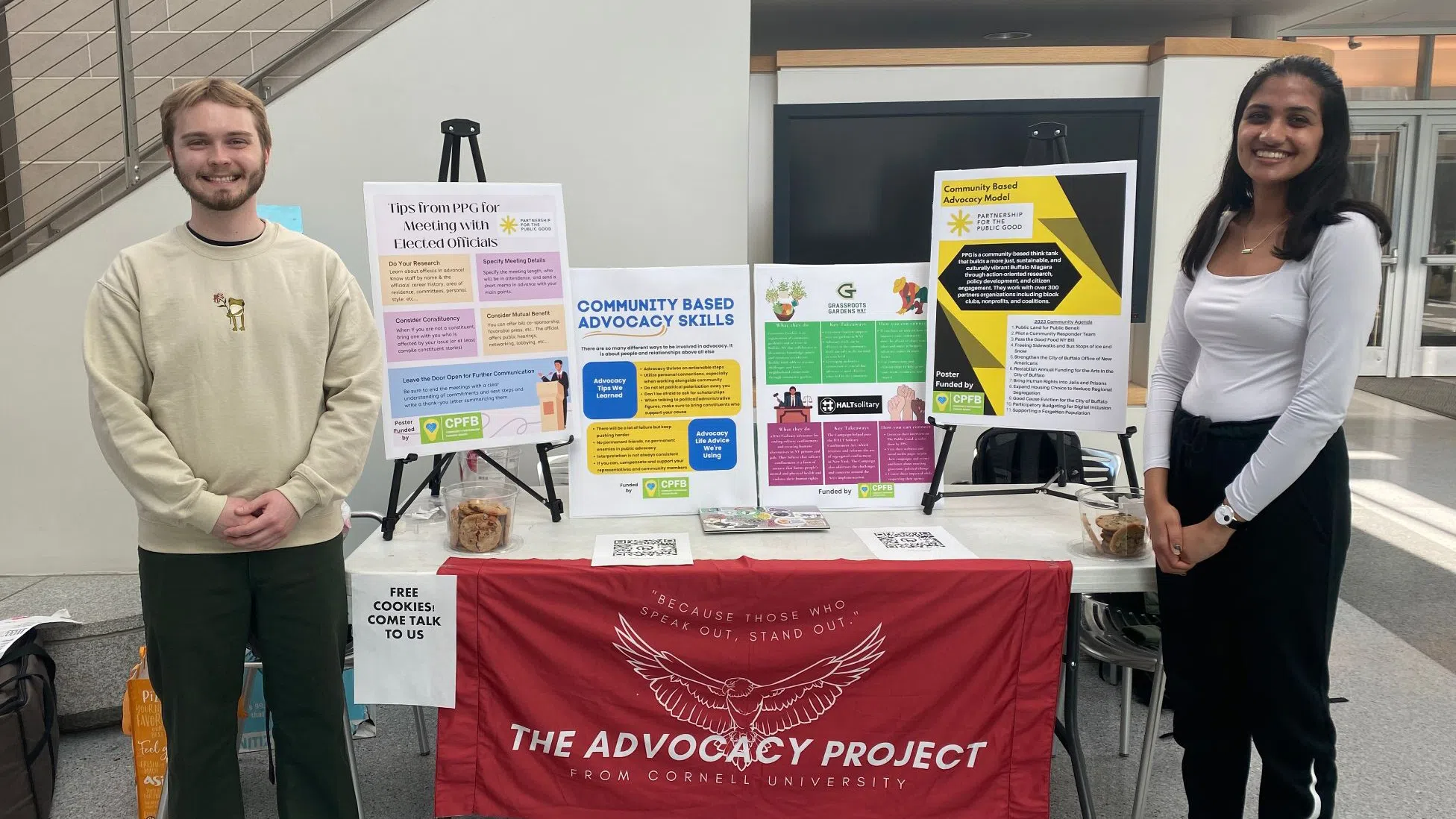 Two college students stand at either side of a table. A red banner in between them reads “The Advocacy Project from Cornell University.” On top of the table, four posters are displayed, and smaller handouts are placed on the table. 