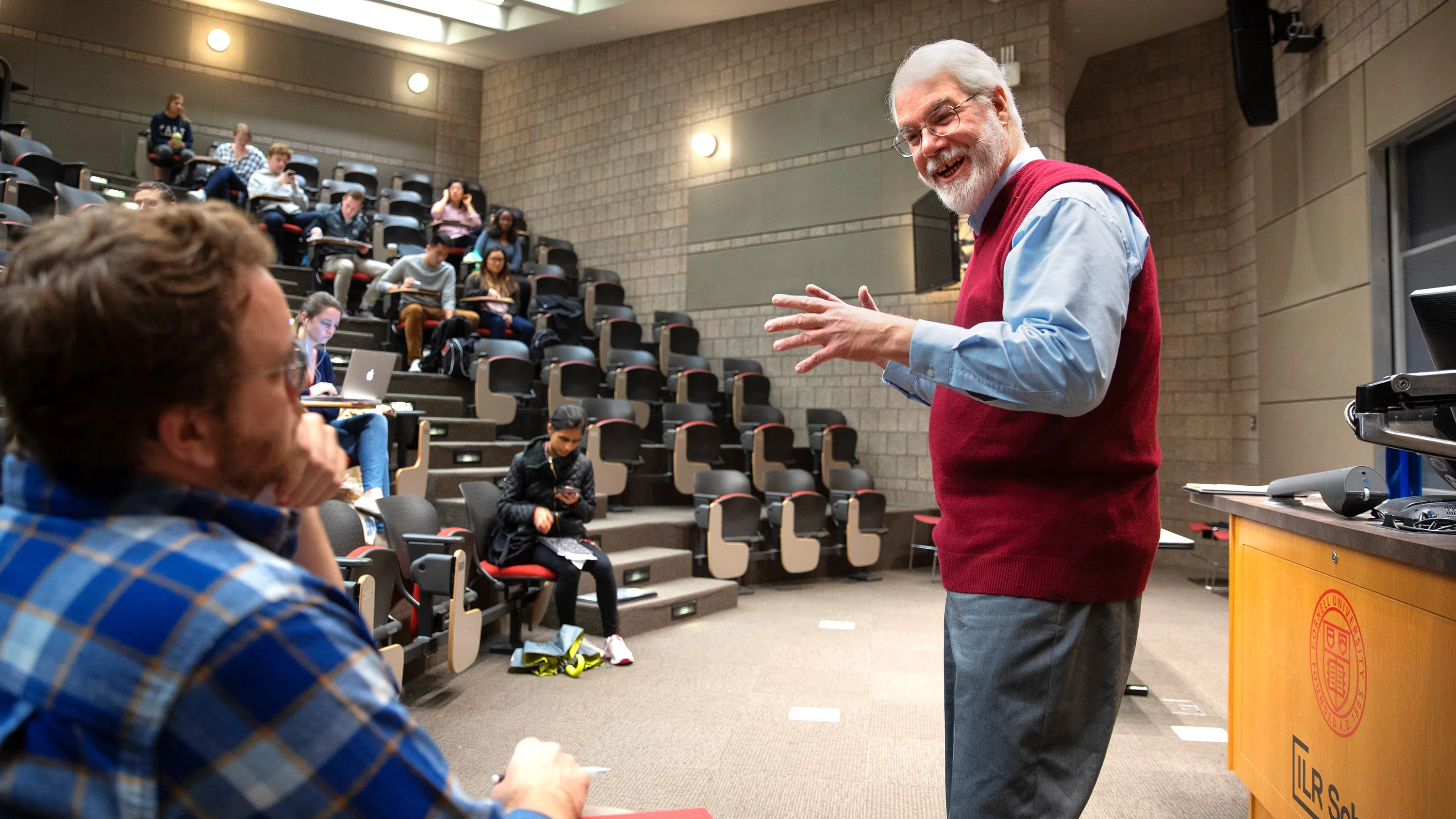 A professor in a lecture hall, hands outstretched, speaks to a student.